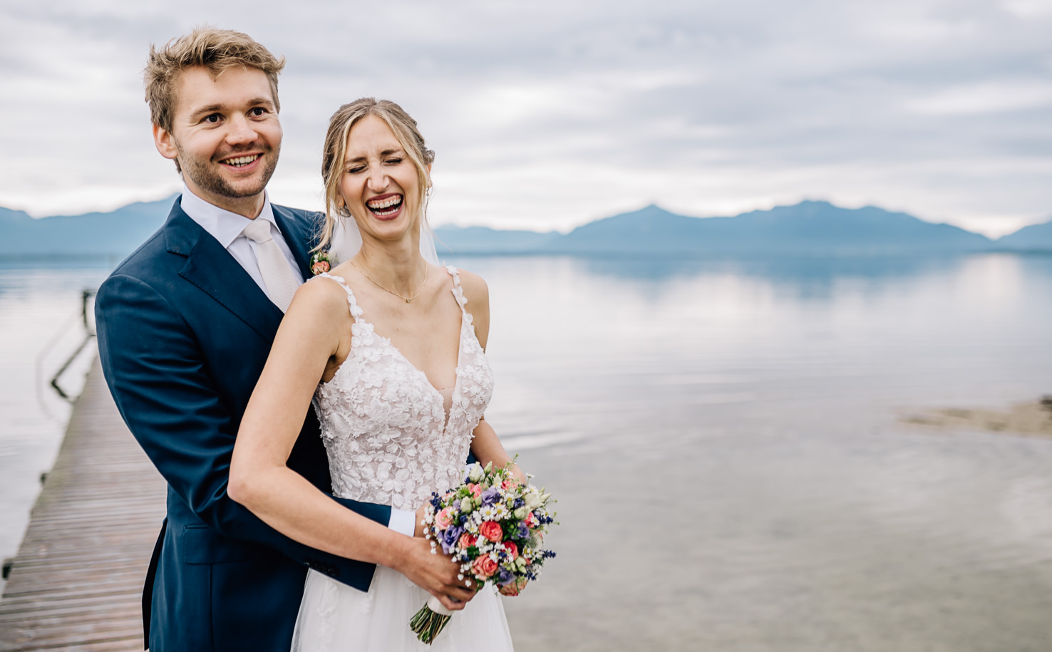 Dieses romantische Hochzeitsfoto von Mica Zeitz zeigt ein glückliches Brautpaar am Ufer eines malerischen Sees, umgeben von sanften Bergen und einem stimmungsvollen Himmel. Die entspannte Atmosphäre und die Liebe, die zwischen den beiden strahlt, machen dieses Bild zu einem perfekten Beispiel für Hochzeitsfotografie.