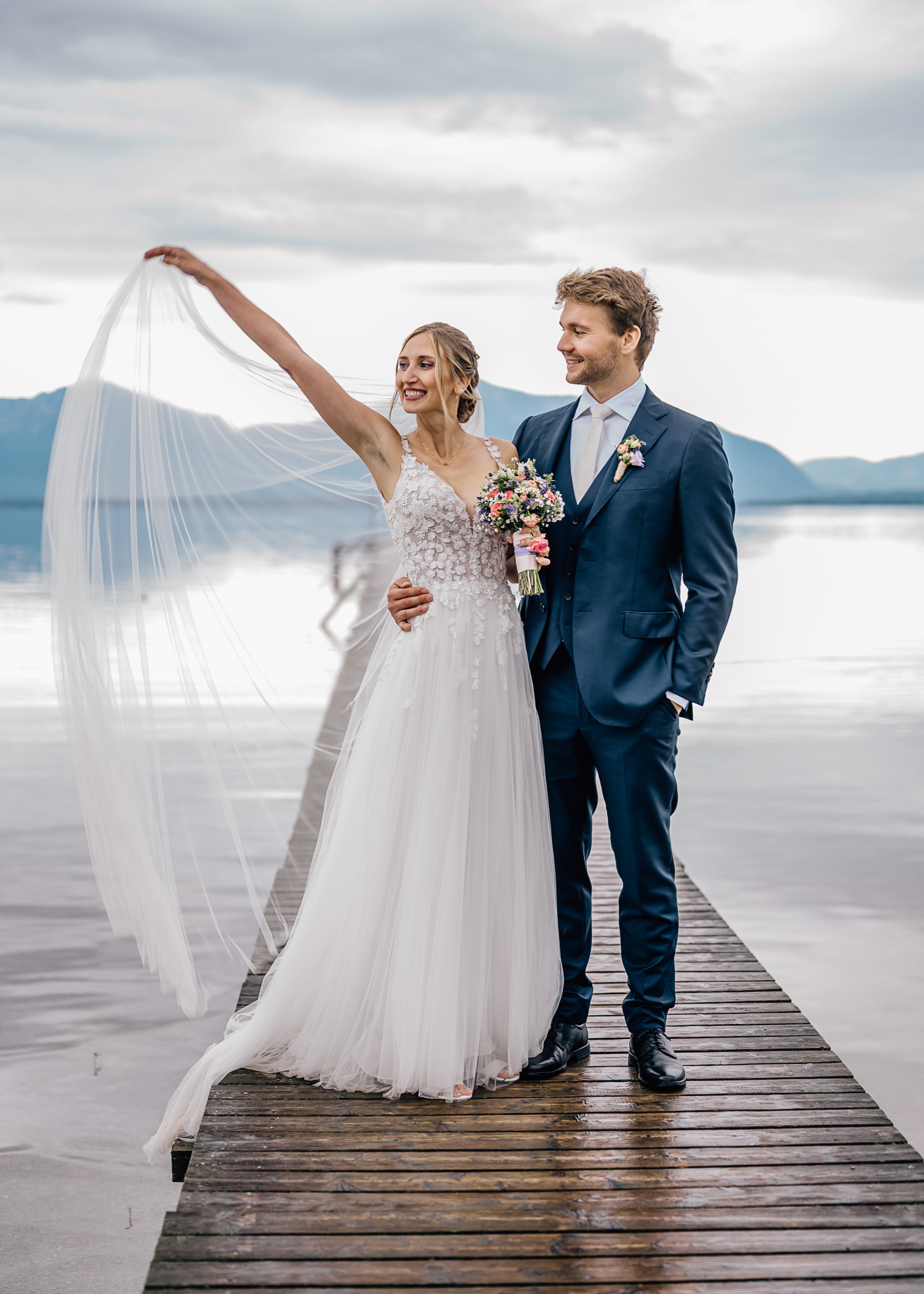 Dieses romantische Hochzeitsbild von Mica Zeitz zeigt ein glückliches Brautpaar auf einem Holzsteg, umgeben von einer malerischen Landschaft. Die Braut in einem zarten, mit Blumen geschmückten Kleid strahlt Freude aus, während der Bräutigam stolz an ihrer Seite steht und die Umgebung eine traumhafte Kulisse für diese unvergessliche Zeremonie bietet.