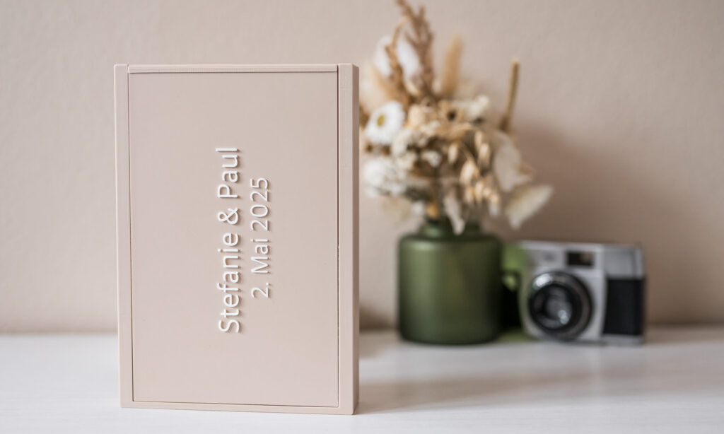Das stilvolle Foto von Mica Zeitz zeigt eine elegante, personalisierte Box für Hochzeitsfotos, die mit den Namen Stefanie und Paul sowie dem Datum 2. Mai 2025 verziert ist. Im Hintergrund sind sanfte Set-Elemente und eine nostalgische Kamera zu sehen, die der Komposition einen besonderen Charme verleihen.
