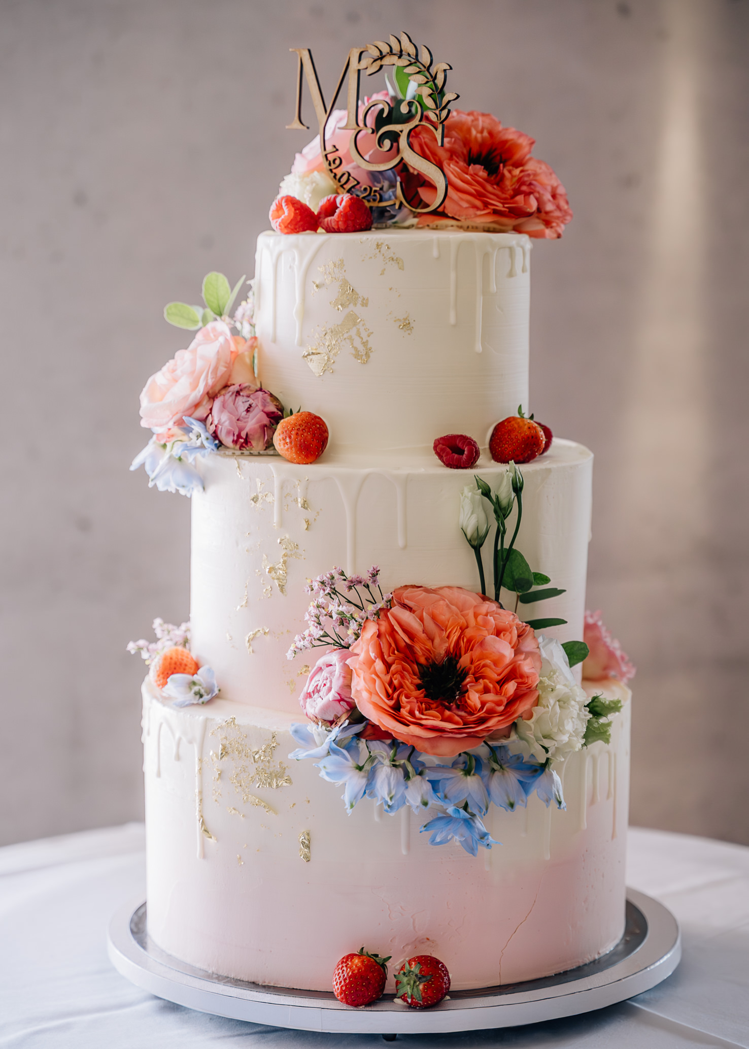 Diese atemberaubende Hochzeitstorte von Mica Zeitz besticht durch ihre mehrstöckige Gestaltung, verziert mit zarten Blüten und frischen Früchten. Mit goldenen Akzenten und einer eleganten Drip-Optik ist sie das perfekte Highlight für jede festliche Feier.