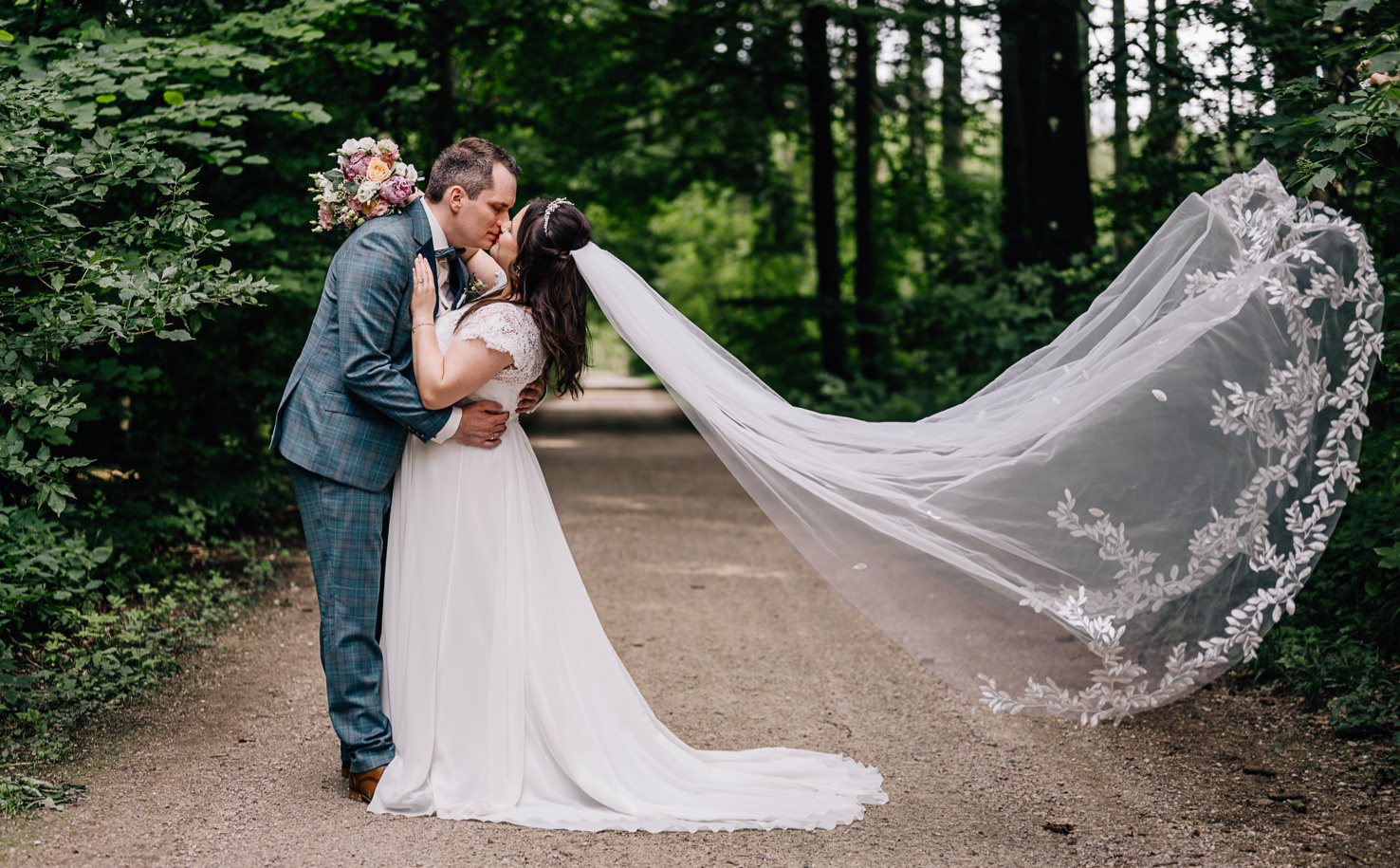 Dieses romantische Hochzeitsbild, festgehalten von Fotografin Mica Zeitz, zeigt ein verliebtes Paar in einer malerischen Waldlandschaft, umgeben von üppigem Grün. Die elegante Braut mit ihrem fließenden Kleid und dem kunstvollen Schleier schafft eine traumhafte Atmosphäre voller Zärtlichkeit und Liebe.
