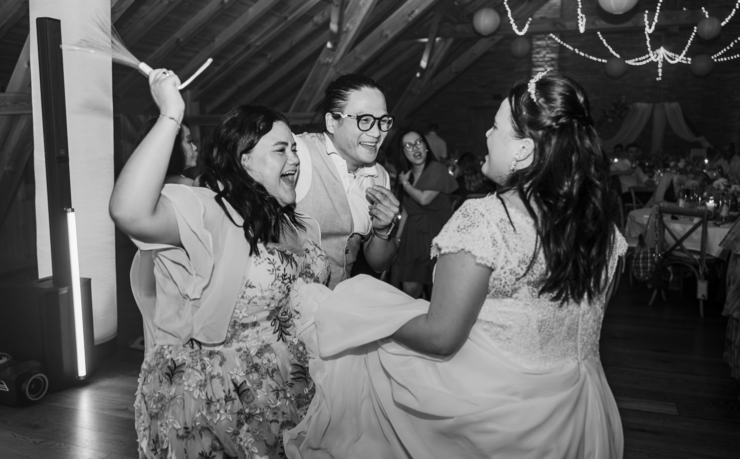 Das lebendige Bild von Mica Zeitz fängt die Freude und das Feiern bei einer Hochzeit in einem rustikalen Ambiente ein, wo Freunde und Familie ausgelassen tanzen und lachen. Die schwarz-weiße Fotografie vermittelt eine nostalgische Stimmung und zeigt die unvergesslichen Momente eines besonderen Tages.
