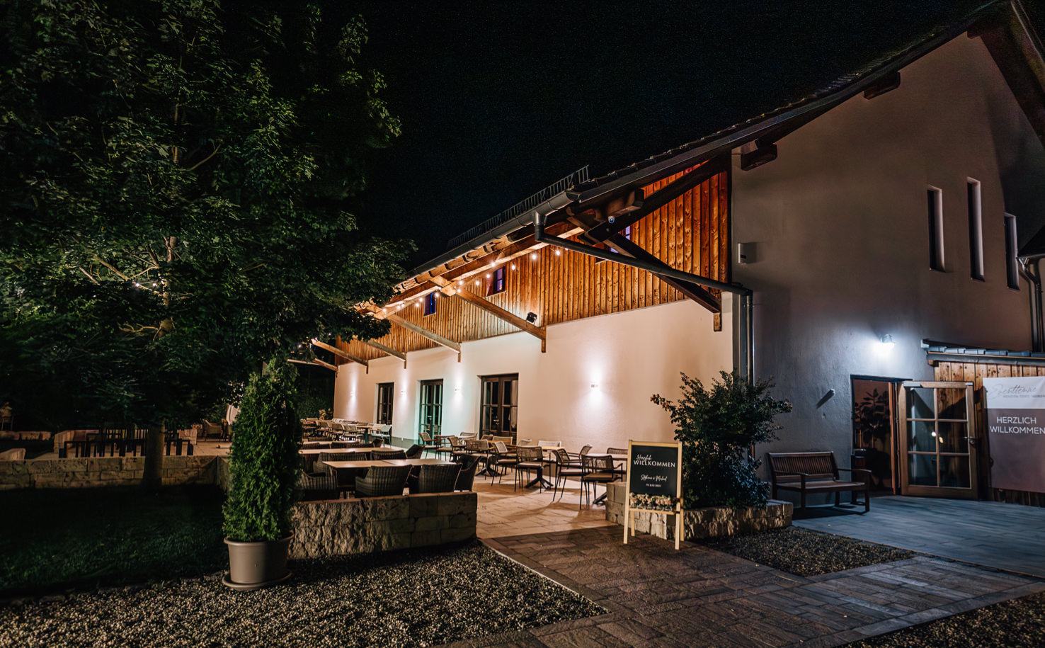 Das stimmungsvolle Nachtbild von Mica Zeitz zeigt ein einladendes Restaurant mit einem gemütlichen Außenbereich, perfekt für romantische Abende oder entspannte Treffen. Die warmen Lichter und die harmonische Architektur schaffen eine einladende Atmosphäre, die Gäste anzieht und zum Verweilen einlädt.