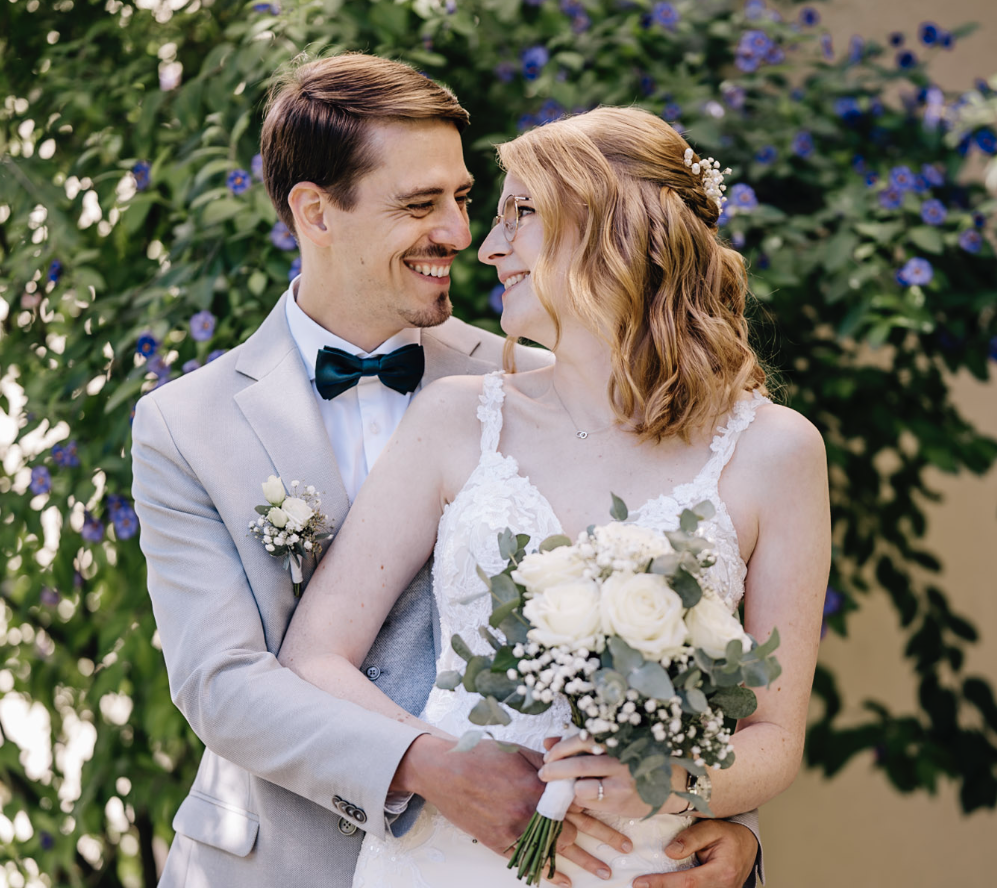 Dieses romantische Hochzeitsbild, aufgenommen von der talentierten Fotografin Mica Zeitz, zeigt ein verliebtes Paar inmitten einer blühenden Kulisse. Die Freude und Nähe der beiden werden durch den strahlenden Blick und den eleganten Brautstrauß aus weißen Rosen und Eukalyptus perfekt unterstrichen.