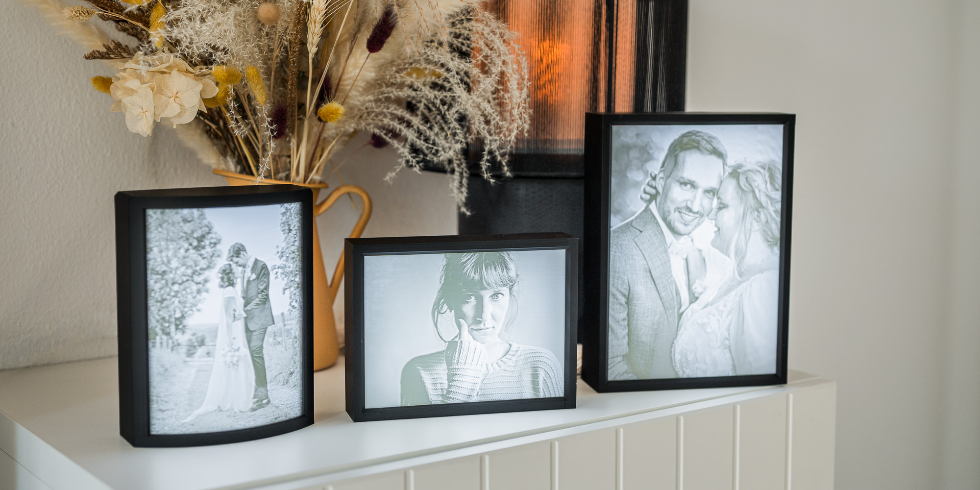 Das Bild zeigt eine stilvolle Anordnung von gerahmten Schwarzweißfotos, die besondere Momente festhalten, ergänzt durch eine ansprechende Trockenblumen-Dekoration. Fotografiert von Mica Zeitz strahlen die Portraits eine zeitlose Eleganz aus und verleihen jedem Raum eine persönliche Note.