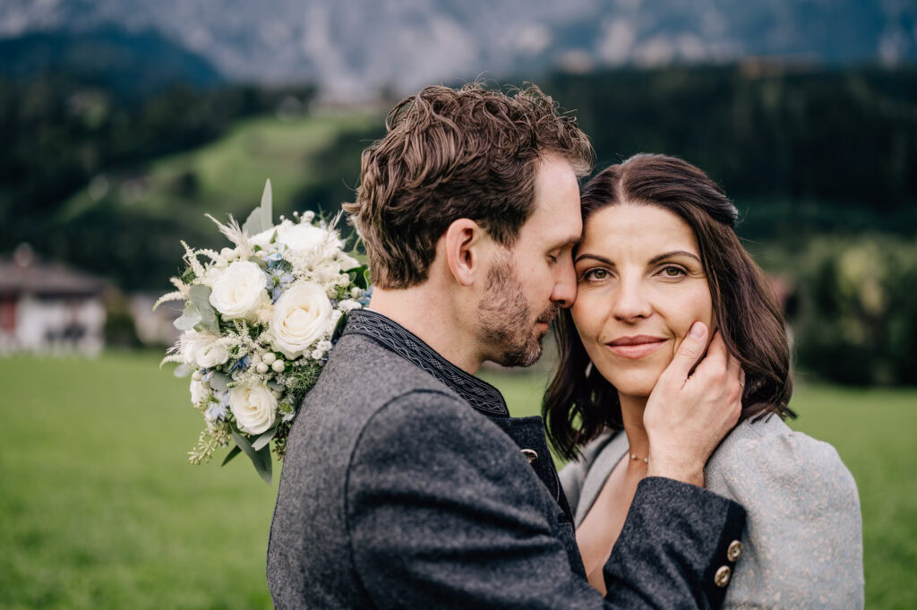 In dieser romantischen Hochzeitsaufnahme, festgehalten von der talentierten Fotografin Mica Zeitz, umarmen sich ein Brautpaar vor einer malerischen Berglandschaft, was die tiefe Verbundenheit und Freude des Augenblicks unterstreicht. Die liebevollen Details des Brautstraußes und die emotionale Nähe der beiden Protagonisten schaffen eine unvergessliche Atmosphäre für jedes Hochzeitsalbum.