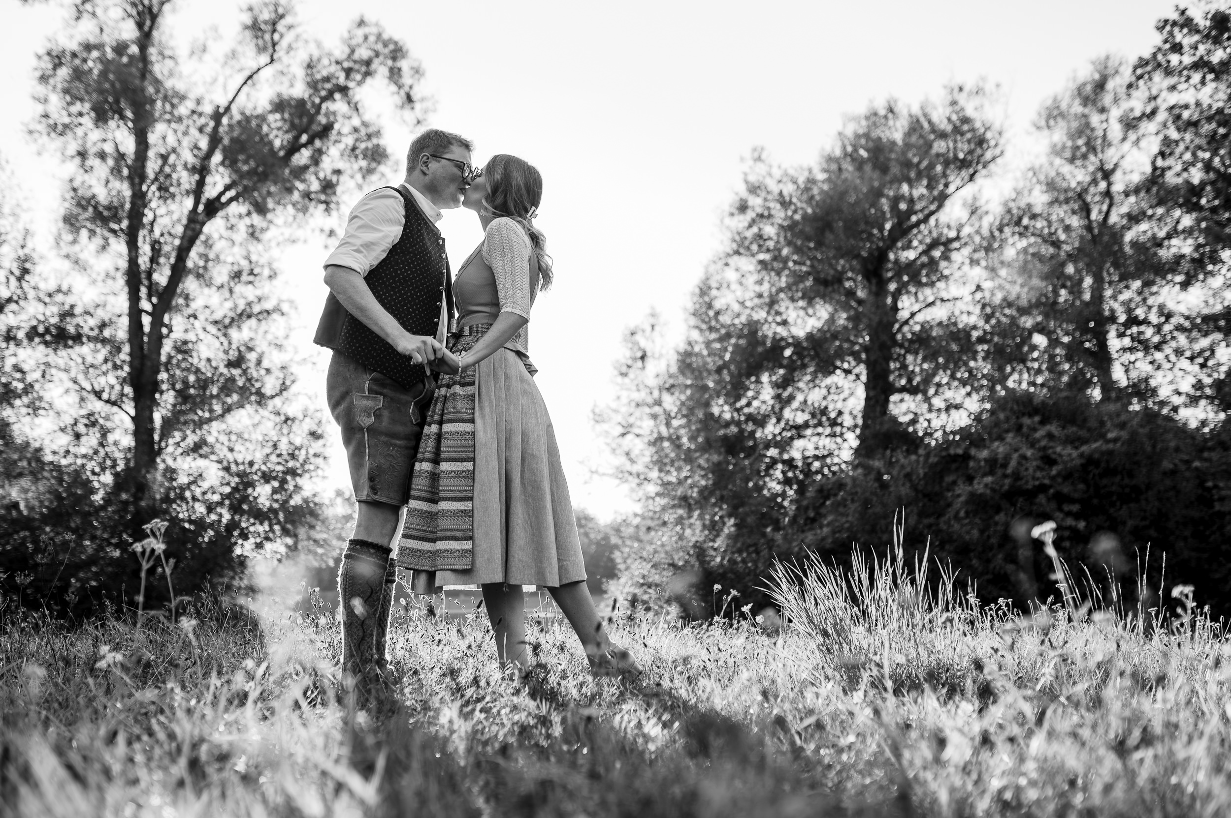 In dieser romantischen schwarz-weiß Aufnahme von Mica Zeitz umarmen sich ein Paar in traditioneller Tracht, während sie sich zärtlich küssen und die unberührte Natur im Hintergrund die Liebe symbolisiert. Die Bildkomposition fängt die intimen Momente der Zuneigung und die Schönheit des ländlichen Lebens perfekt ein.