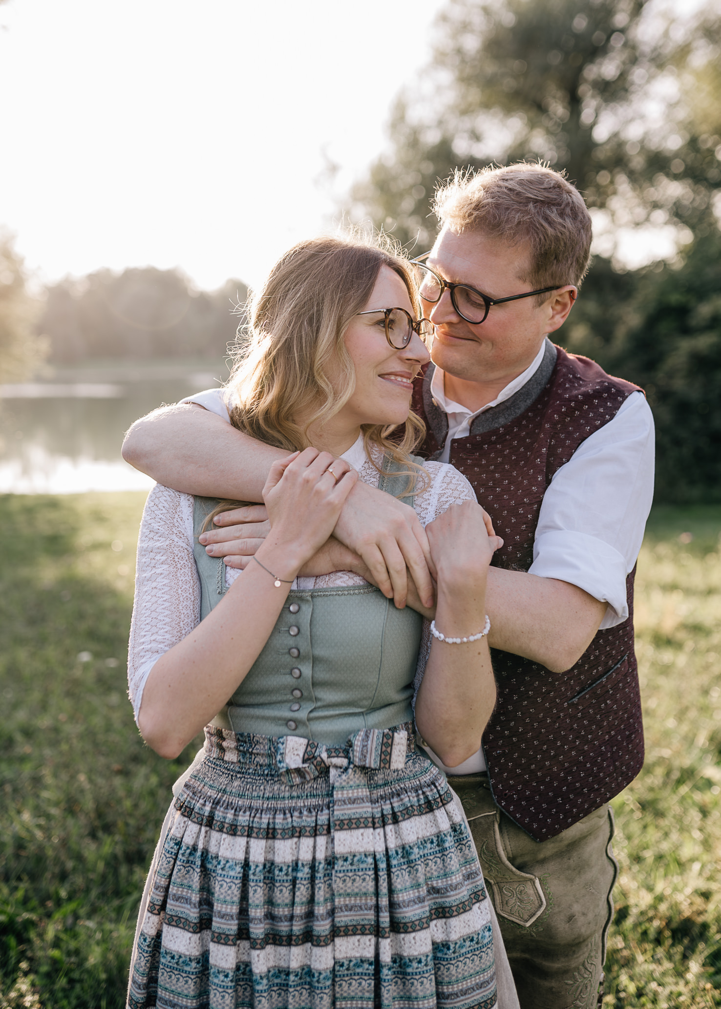 In diesem romantischen Paarporträt, aufgenommen von der Fotografin Mica Zeitz, umarmen sich die Liebenden sanft vor einer malerischen Kulisse mit sanftem Licht und grüner Natur. Die traditionelle Tracht verleiht diesem Moment eine besondere bayerische Note, perfekt für Hochzeits- und Verlobungsfotografie.