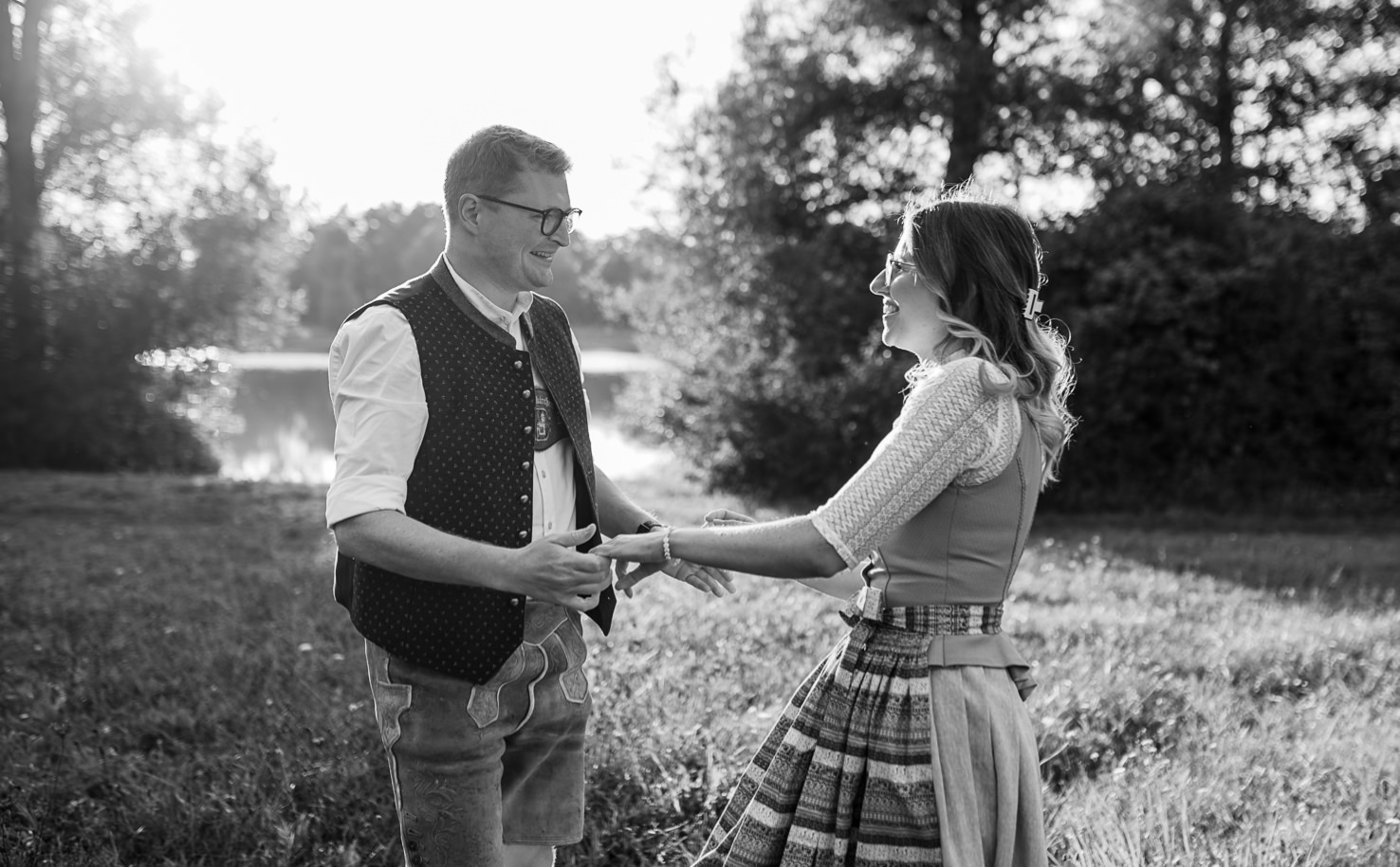 Auf diesem romantischen Schwarz-Weiß-Foto von der Fotografin Mica Zeitz lächeln ein Paar verliebt und hält sich an den Händen, während sie in einer malerischen Freiluftkulisse stehen. Die traditionelle Tracht des Paares fügt dem Bild einen charmanten, kulturellen Flair hinzu, ideal für Hochzeitsfotografie und Paarkunst.