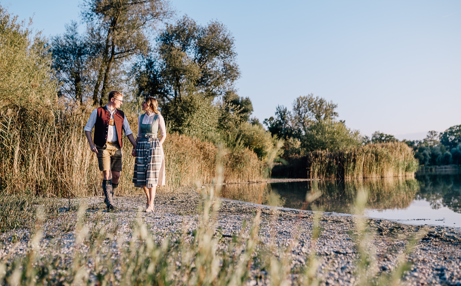 In dieser malerischen Szene, festgehalten von der Fotografin Mica Zeitz, spazieren ein Paar in traditioneller bayerischer Tracht entlang eines friedlichen Gewässers, umgeben von üppiger Natur. Die harmonische Verbindung von Mensch und Landschaft unterstreicht die Schönheit traditioneller Trachten und die idyllische Atmosphäre des ländlichen Raums.