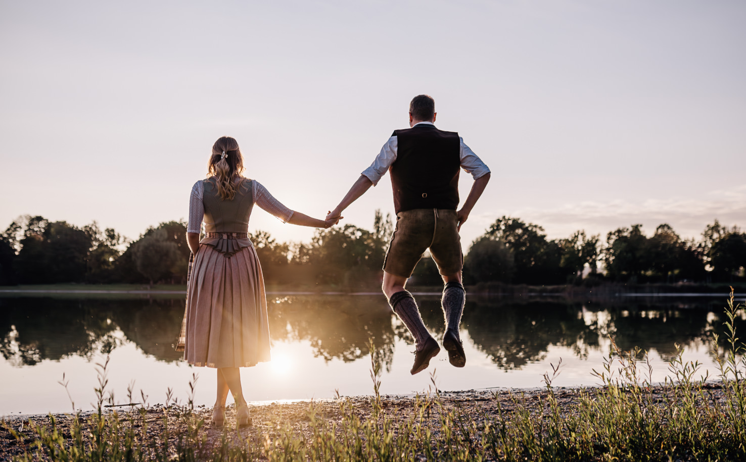 In diesem romantischen Bild von Mica Zeitz feiern ein Paar in traditioneller Tracht einen unvergesslichen Moment am idyllischen Wasser. Der Sonnenuntergang spiegelt sich in der Oberfläche und verstärkt die emotionale Verbindung der beiden, während sie Hand in Hand in die Zukunft springen.