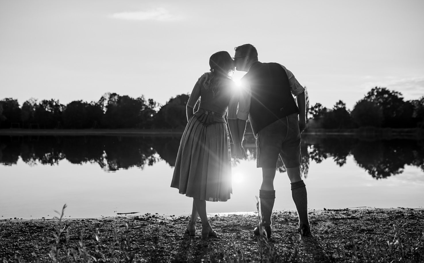 Das eindrucksvolle Schwarz-Weiß-Foto von Mica Zeitz zeigt ein romantisches Paar, das sich am Ufer eines ruhigen Sees küsst, während die Sonne im Hintergrund strahlt. Diese stimmungsvolle Aufnahme vermittelt Liebe und Verbundenheit in einer malerischen Naturkulisse und eignet sich perfekt für Hochzeits- und Verlobungsfotografie.