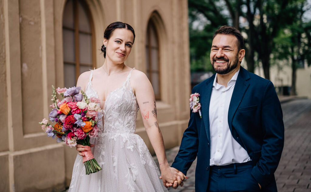 Das Bild zeigt ein fröhliches Brautpaar, das Händchen haltend in einer malerischen Umgebung in Ingolstadt spaziert. Die Fotografin Mica Zeitz hat diesen besonderen Moment in beeindruckender Weise eingefangen, wobei die strahlenden Gesichter des Paares und der farbenfrohe Brautstrauß im Fokus stehen.