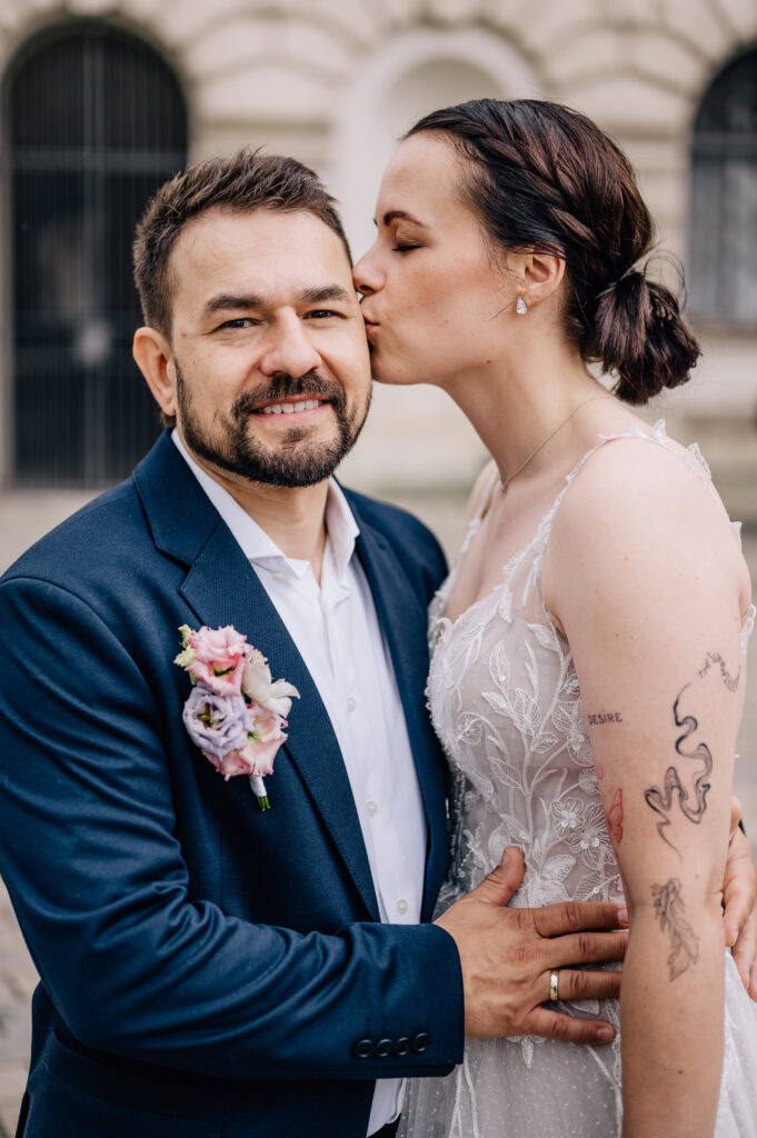 Das Bild zeigt ein liebevolles Paar, das sich in der Ingolstädter Innenstadt gekonnt für ein Hochzeitsfoto posiert, während die Braut ihrem Partner zärtlich einen Kuss auf die Wange gibt. Dieses emotionale Moment wurde von der Fotografin Mica Zeitz festgehalten, und die Umgebung verleiht der Bildkomposition zusätzlichen Charme.