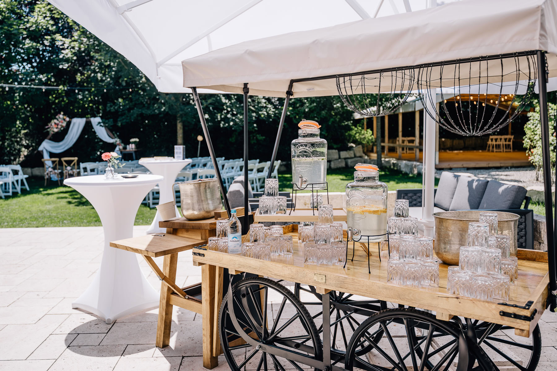 Das Bild zeigt eine einladende Hochzeitslocation im Eventtenne Pfaffenhofen mit einem stilvollen Getränkewagen, der charmant dekoriert ist. Die ansprechende Gartenumgebung und die eleganten Stehtische bieten eine perfekte Kulisse für unvergessliche Feierlichkeiten, was diese Location zur Empfehlung macht. Fotografin Mica Zeitz.