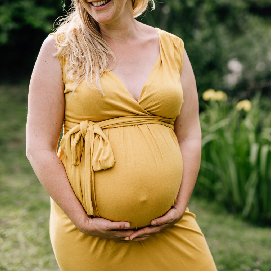 Das Bild zeigt eine glücklich lächelnde Frau in einem eleganten, gelben Leihoutfit, die sanft ihren Babybauch hält. Die Aufnahme von Mica Zeitz fängt die Schönheit der Schwangerschaft in einer natürlichen Umgebung ein.