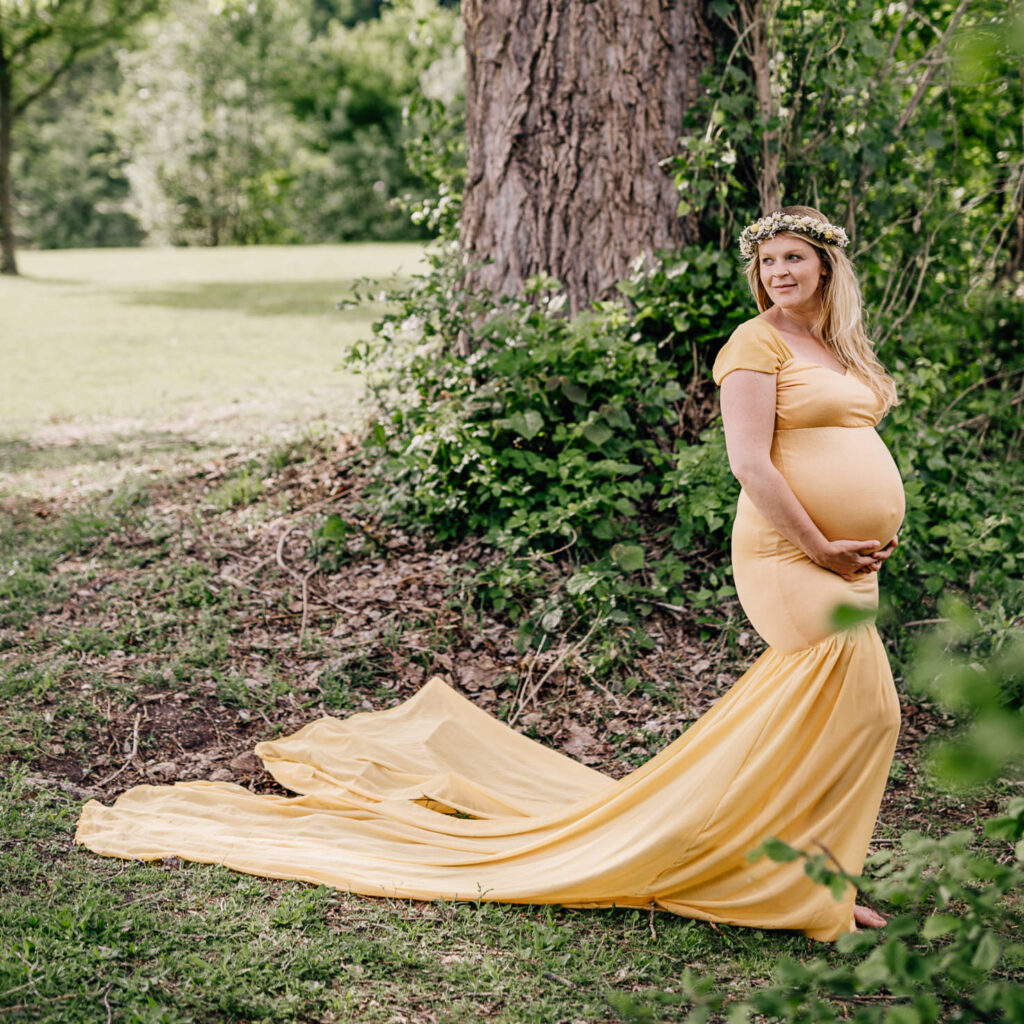 Das Bild, aufgenommen von Mica Zeitz, zeigt eine schwangere Frau in einem eleganten Leihoutfit mit einem langen, fließenden Saum, der sanft über den Boden gleitet. Umgeben von der Natur strahlt sie mit einem sanften Lächeln und einem Blumenkranz im Haar, was die Schönheit der Babybauchfotos unterstreicht.
