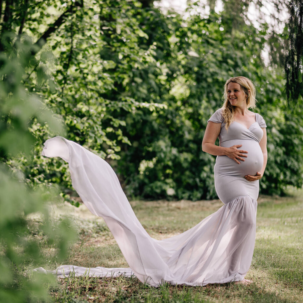 Das Bild zeigt eine schwangere Frau in einem eleganten Leihoutfit, das mit einem fließenden Stoff versehen ist, der sanft im Wind weht. Die Aufnahme, die von der Fotografin Mica Zeitz gemacht wurde, fängt die Schönheit von Babybauchfotos in einer grünen, naturnahen Umgebung ein.