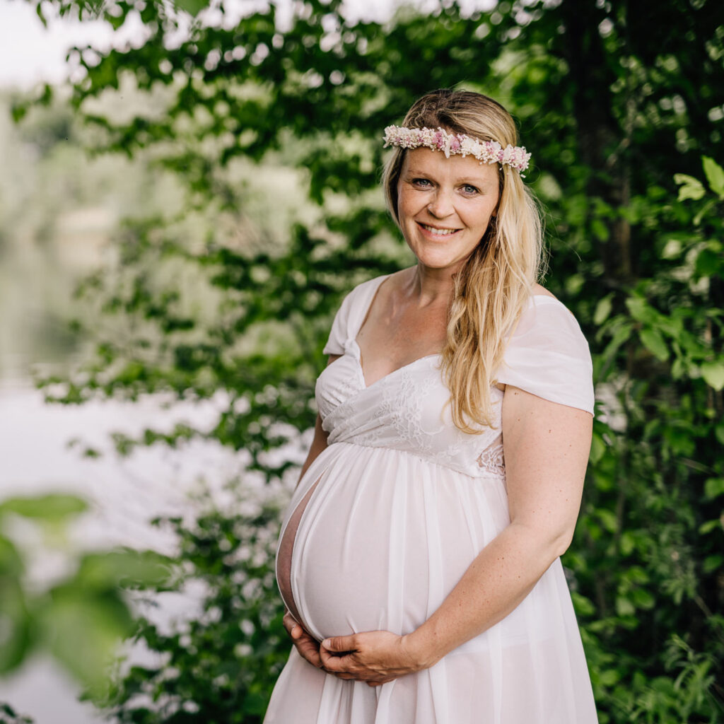 Das Bild zeigt eine werdende Mutter in einem eleganten Leihoutfit, die stolz ihren Babybauch präsentiert. Umrahmt von üppigem Grün strahlt sie eine natürliche Schönheit und Freude aus, festgehalten von der Fotografin Mica Zeitz.