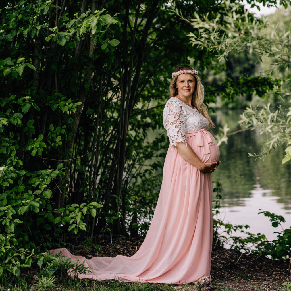 Das Bild zeigt eine schwangere Frau in einem eleganten Leihoutfit, das aus einem zarten, rosa Kleid mit Spitzenärmel besteht, umgeben von üppigem Grün am Ufer eines Gewässers. Diese Babybauchfotos wurden von der Fotografin Mica Zeitz aufgenommen und fangen die natürliche Schönheit und Vorfreude der Schwangeren ein.