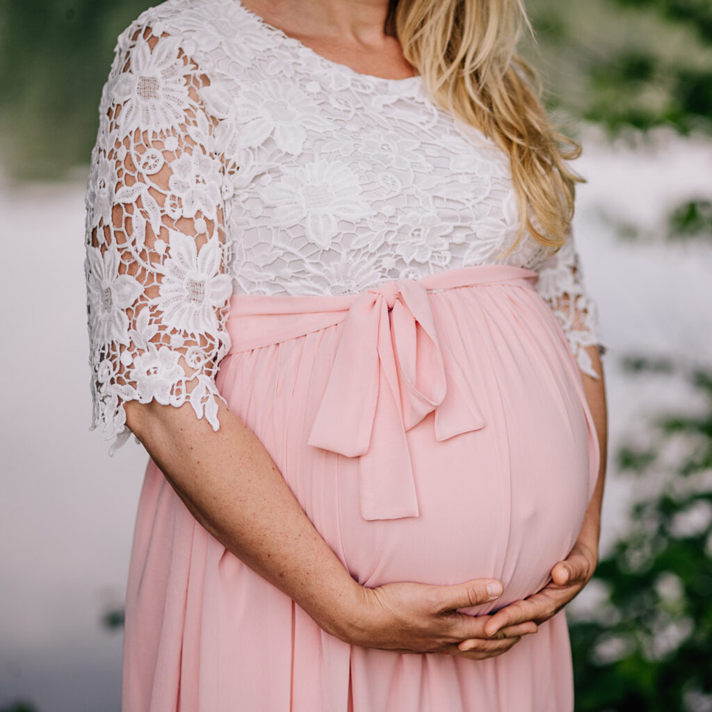 Auf dem Bild von Fotografin Mica Zeitz trägt eine schwangere Frau ein elegantes Leihoutfit aus zarter Spitze und einem rosa Rock. Sie hält liebevoll ihren Babybauch, was die Vorfreude auf das Neugeborene symbolisiert.
