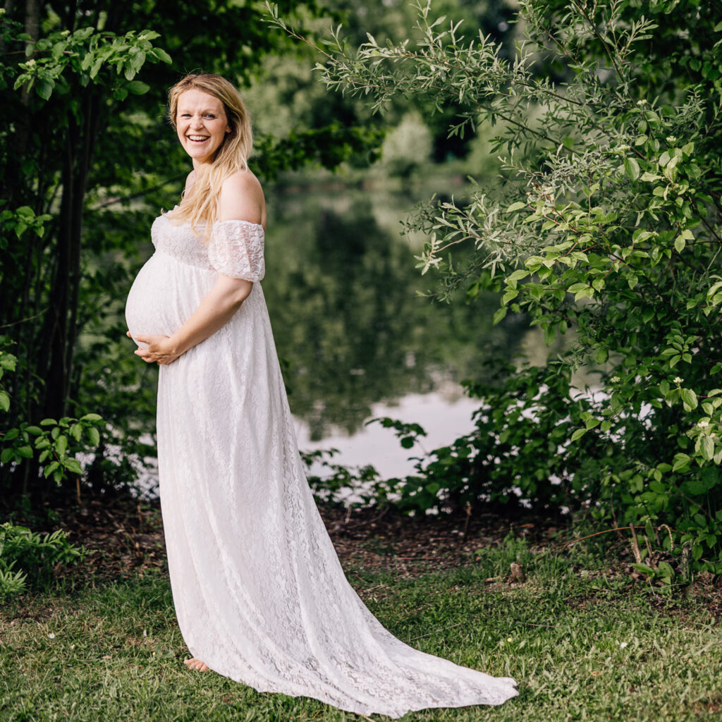Auf dem Bild ist eine werdende Mutter in einem eleganten Leihoutfit zu sehen, die glücklich ihren Babybauch hält. Die malerische Kulisse mit Wasser und üppigem Grün unterstreicht die Freude und das Glück der Schwangerschaft, festgehalten von der Fotografin Mica Zeitz.