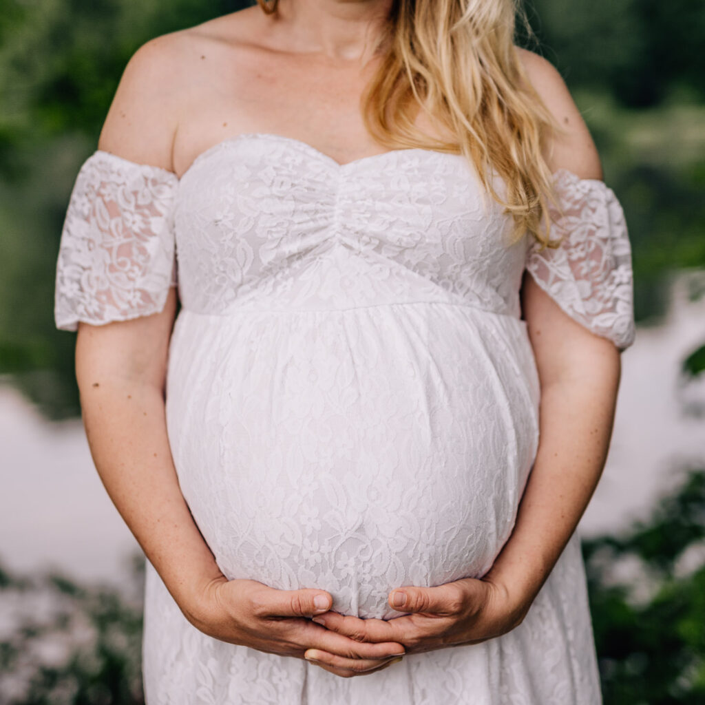 Das Bild zeigt eine schwangere Frau in einem eleganten Leihoutfit mit Spitzenärmeln, die liebevoll ihren Babybauch umarmt. Die Aufnahme von Mica Zeitz fängt die Vorfreude und Schönheit der Schwangerschaft in einer natürlichen Umgebung ein.