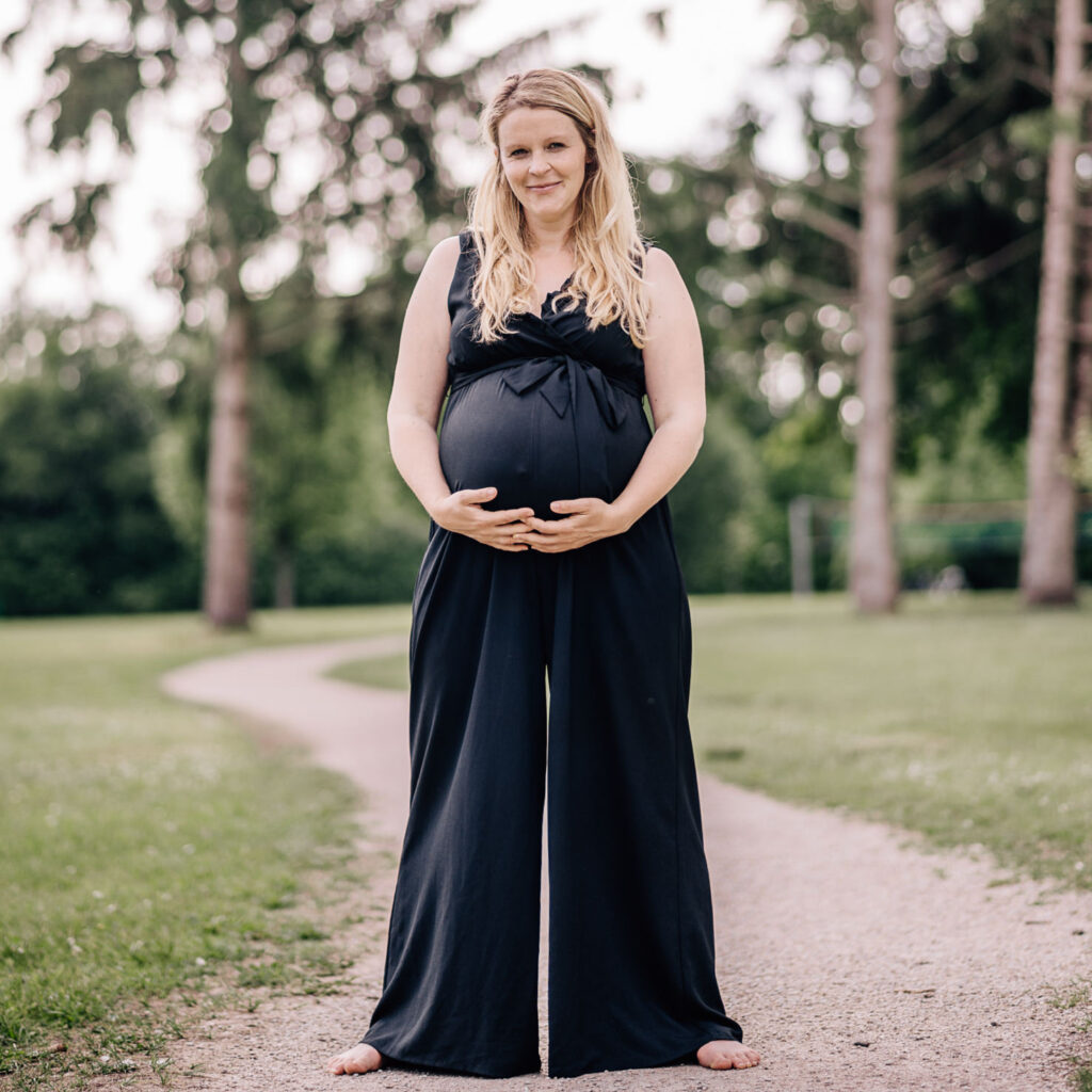 Das Bild zeigt eine schwangere Frau in einem eleganten Leihoutfit, die sanft ihren Babybauch hält. Die Aufnahme, die von der Fotografin Mica Zeitz stammt, vermittelt eine ruhige und natürliche Atmosphäre in einem parkähnlichen Umfeld.