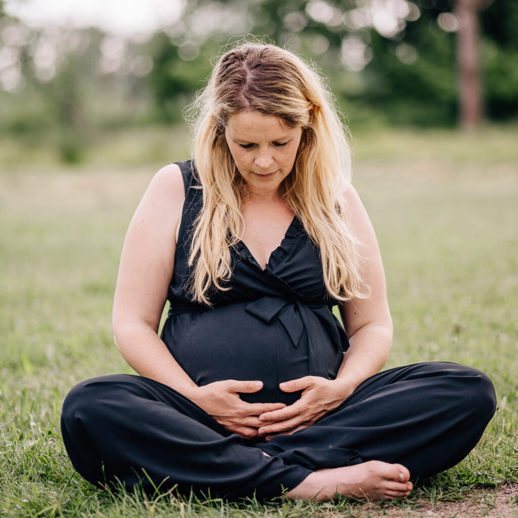 Das Bild zeigt eine schwangere Frau in einem eleganten Leihoutfit, die liebevoll ihren Babybauch betrachtet. Die natürliche Umgebung und die gelassene Pose verleihen der Aufnahme eine sanfte, emotionale Atmosphäre, festgehalten von der Fotografin Mica Zeitz.
