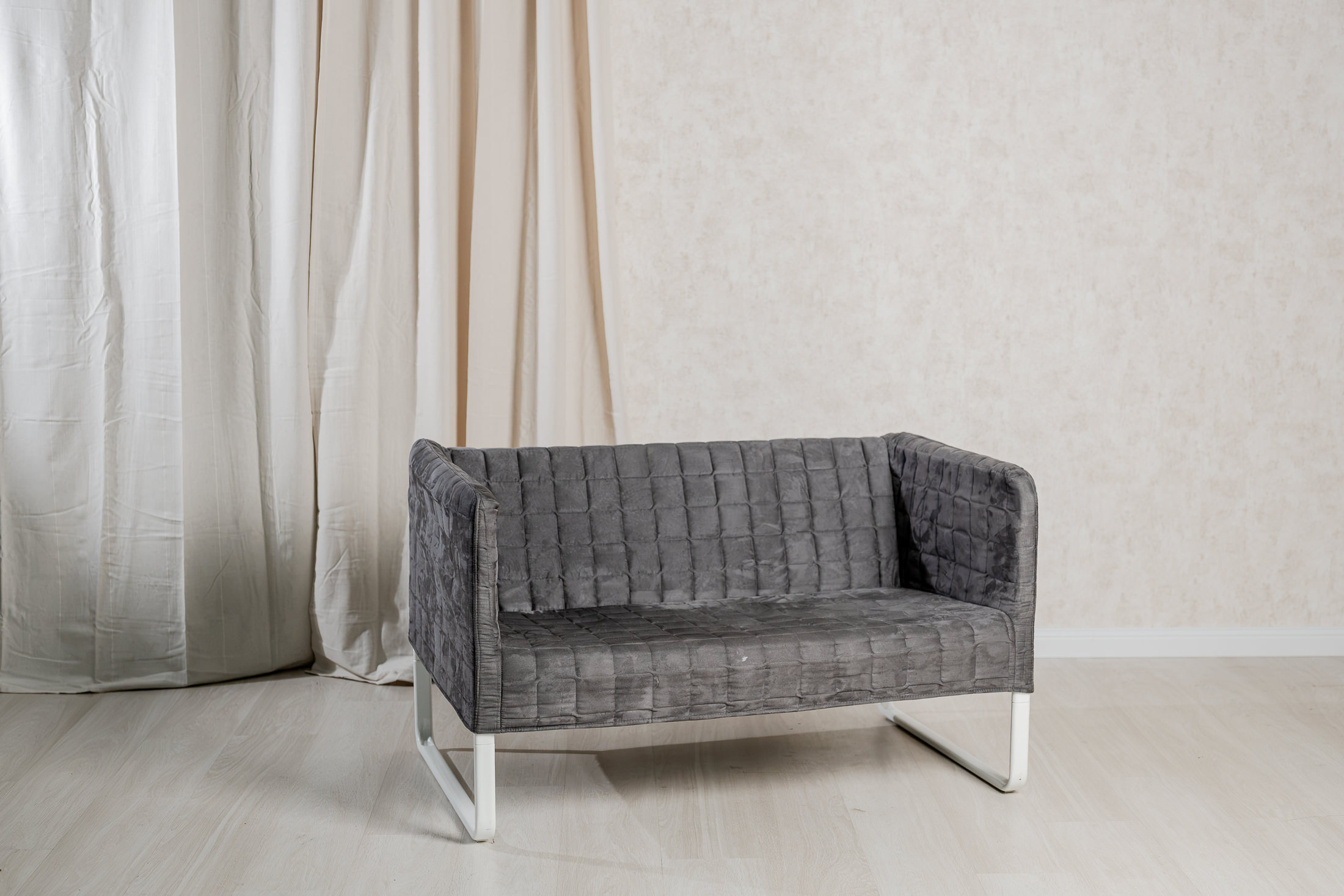 Das Bild zeigt ein modernes, graues Sofa mit einer gesteppten Oberfläche, das in einem hellen Fotostudio in Baar-Ebenhausen steht. Der minimalistische Stil des Sofas harmoniert gut mit dem neutralen Hintergrund und den weichen Vorhängen, festgehalten von Fotografin Mica Zeitz.