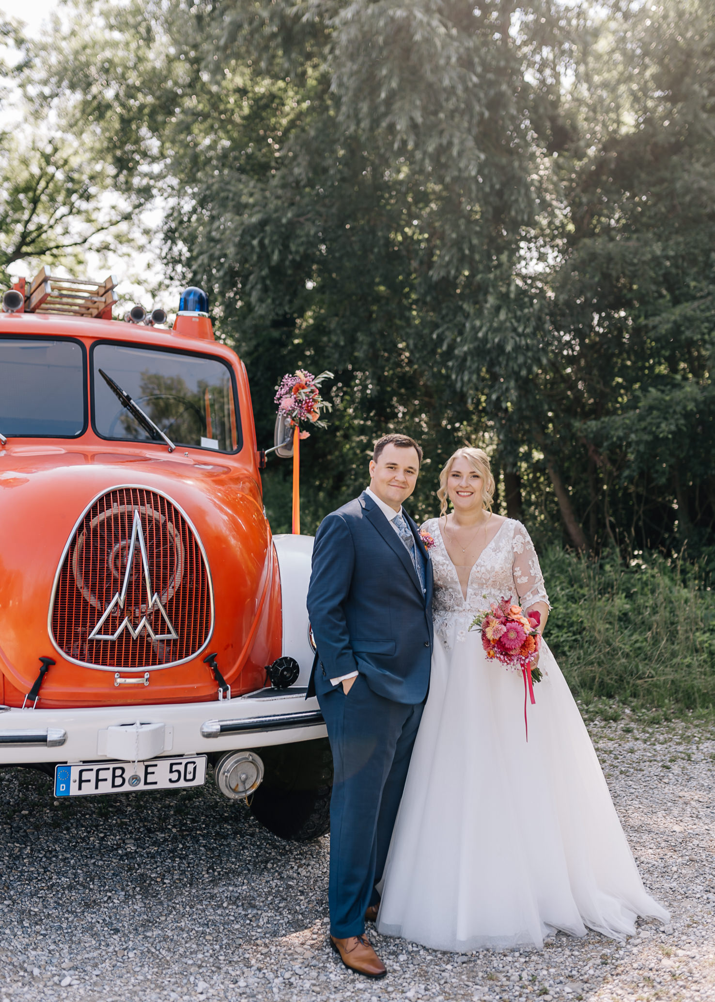 Das Hochzeitsfoto zeigt ein glückliches Paar, das eng vor einem auffälligen roten Feuerwehrauto in Olching steht. Die wunderschöne Braut trägt ein elegantes Kleid und hält einen bunten Blumenstrauß, während der Bräutigam lächelnd an ihrer Seite steht, festgehalten von der Fotografin Mica Zeitz.