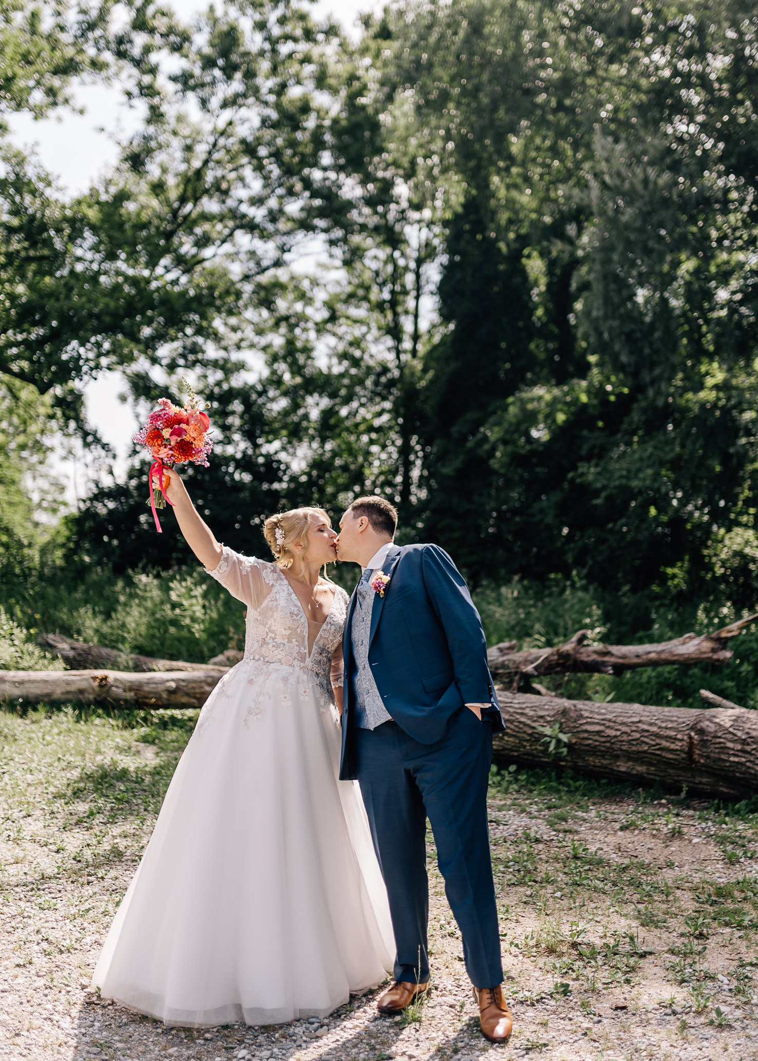 Auf dem Hochzeitsfoto in Olching posieren ein glückliches Paar, während die Braut ihren wunderschönen Blumenstrauß in die Höhe hält. Das Bild strahlt Freude und Romantik aus und wurde von der Fotografin Mica Zeitz aufgenommen.