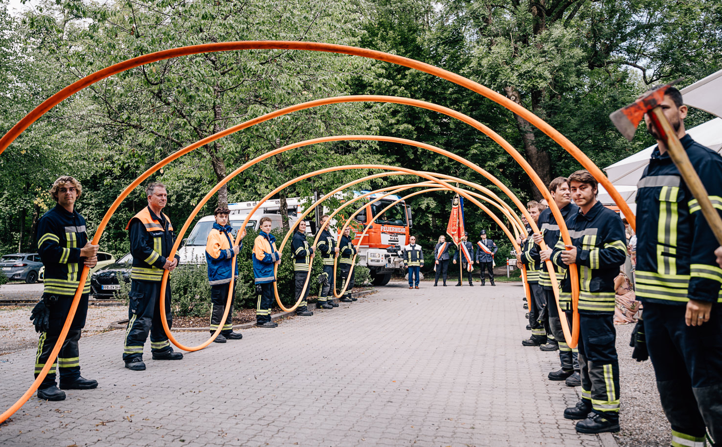 Das Bild zeigt eine Gruppe von Feuerwehrleuten in Olching, die mit orangenen Schläuchen eine Art Tunnel bilden. Es handelt sich um eine feierliche Szene, die eindrucksvoll zur Hochzeitsfeier beiträgt, festgehalten von der Fotografin Mica Zeitz.