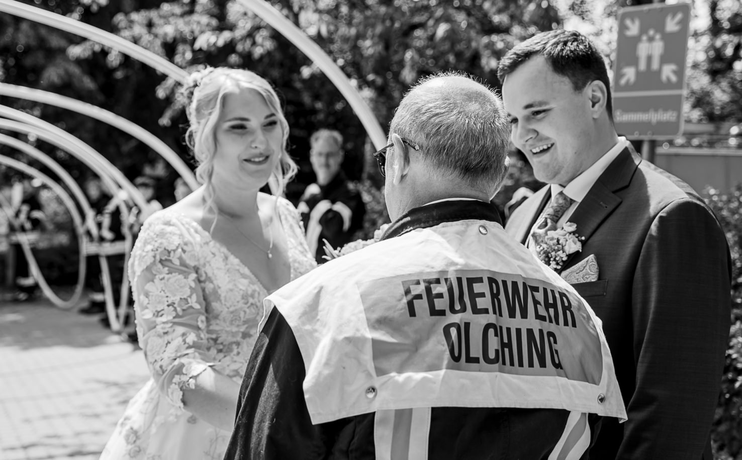 Das Hochzeitsfoto von Mica Zeitz zeigt ein glückliches Brautpaar in Olching, das sich mit einem Feuerwehrmann in Verbindung setzt. Die Szene vermittelt eine herzliche Atmosphäre, während die beiden in eleganten, festlichen Outfits elegant in die Kamera lächeln.