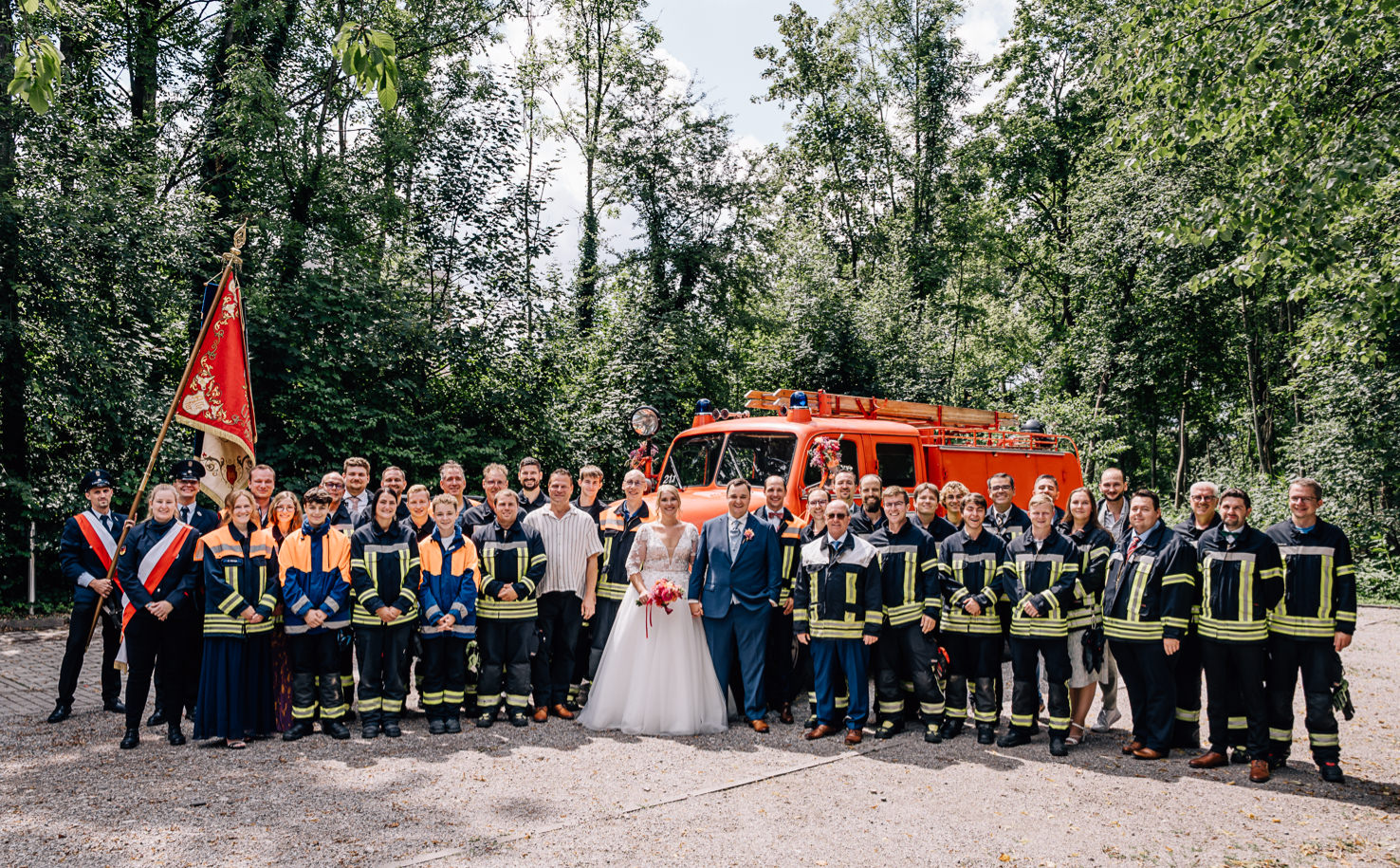 Das Bild zeigt eine fröhliche Hochzeitsgruppe in Olching, bestehend aus dem Brautpaar und zahlreichen Feuerwehrkameraden, die in Uniform vor einem Feuerwehrauto posieren. Fotografiert von Mica Zeitz, vermittelt das Bild eine festliche und gemeinschaftliche Atmosphäre.
