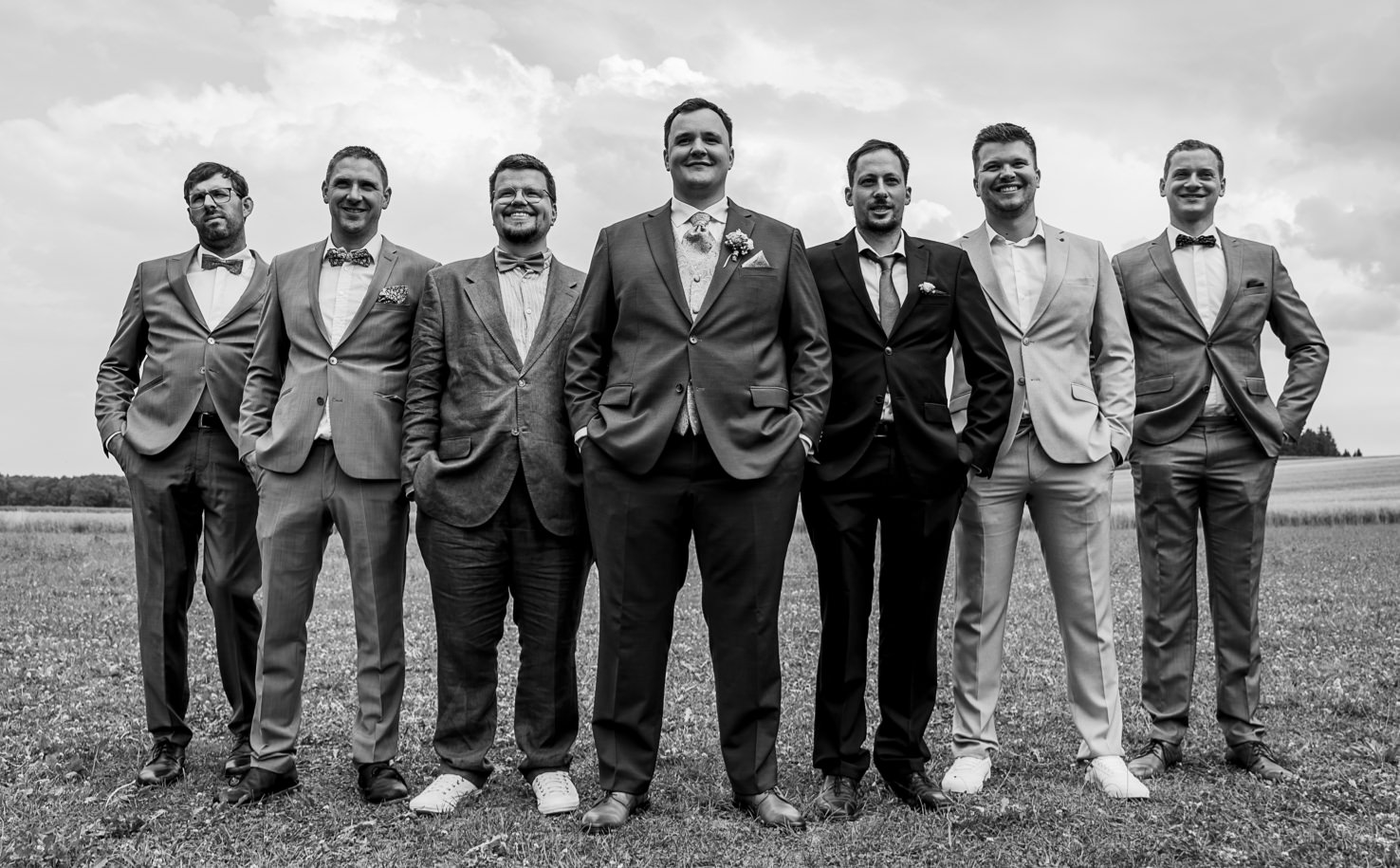 Das Bild, aufgenommen von Mica Zeitz, zeigt eine Gruppe von sieben Männern in eleganter Hochzeitskleidung, die selbstbewusst in einer Reihe stehen. Die Szene ist in einem schönen, offenen Feld in Olching aufgenommen, was die festliche Stimmung der Hochzeitsfeierlichkeit unterstreicht.