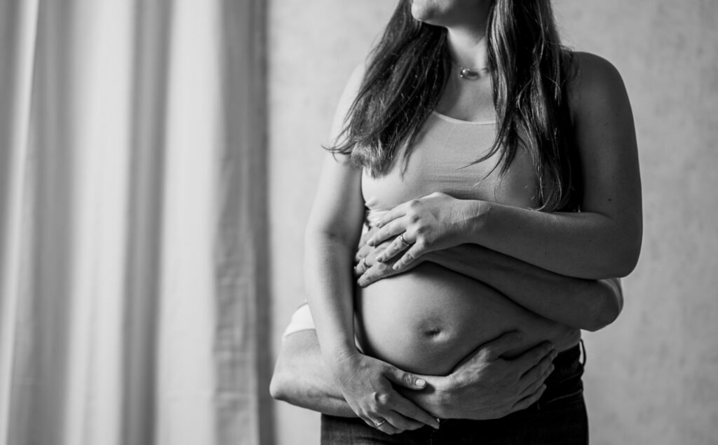 Das Bild zeigt ein emotionales Babybauchfoto, das im GMW-Studio aufgenommen wurde, und fängt einen Moment der Zuneigung zwischen zwei Menschen ein. Die Hände des Partners umarmen liebevoll den Babybauch, was die Vorfreude auf das kommende Baby symbolisiert. Fotografin Mica Zeitz hat diesen besonderen Augenblick wunderbar festgehalten.